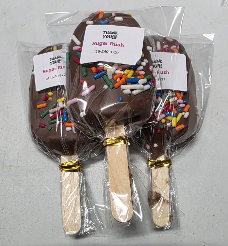 Three chocolate cakesicles with sprinkles and Sugar Rush thank-you labels